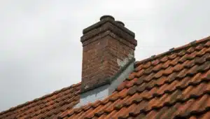 Verweerde bakstenen schoorsteen met uitgebrokkelde voegen en scheefstaande kap op een Nederlands pannendak.