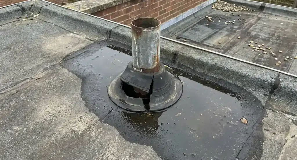 Beschadigde dakdoorvoer met roestige metalen buis op plat dak, omgeven door water en natte membraan, toont lekkage rond de